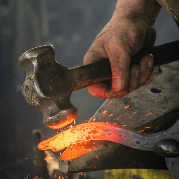 Nahaufnahme einer Handwerker-Hand, die mit einem Hammer glühendes Metall auf einem Amboss bearbeitet – Symbolbild für traditionelles Handwerk.