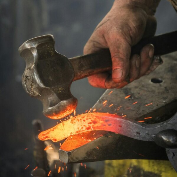 Nahaufnahme einer Handwerker-Hand, die mit einem Hammer glühendes Metall auf einem Amboss bearbeitet – Symbolbild für traditionelles Handwerk.