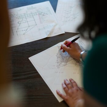 Nahaufnahme eines Ingenieurs, der technische Gerüstbau-Zeichnungen und Statik-Pläne mit einem Stift manuell überprüft und korrigiert.